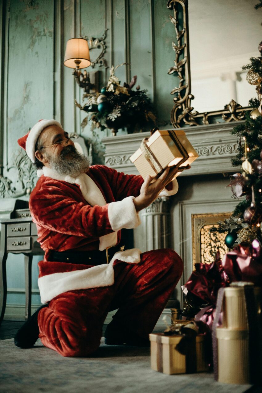 Santa Claus joyfully holding gifts by a decorated Christmas tree indoors.